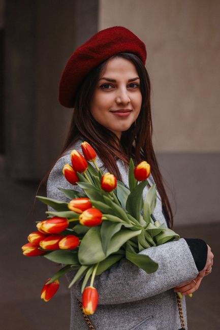 A young woman with long dark hair, wearing a red beret and a grey coat, holds a large bouquet of vibrant orange-red tulips with green leaves in her arms. The tulips are fresh, with smooth petals and s