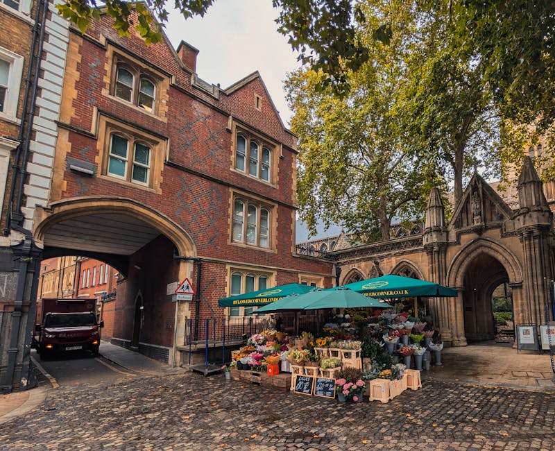 A flower stall positioned on a cobbled street near a historic red-brick building with an arched gateway, featuring a green canopy with the website floristholborn.co.uk. The stall displays a vibrant ar