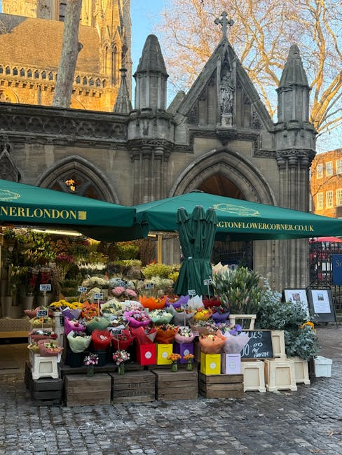 A vibrant flower stall set up outdoors in front of a historic Gothic-style building with pointed arches and decorative stonework in Holborn. The stall is shaded by a large green umbrella with branding