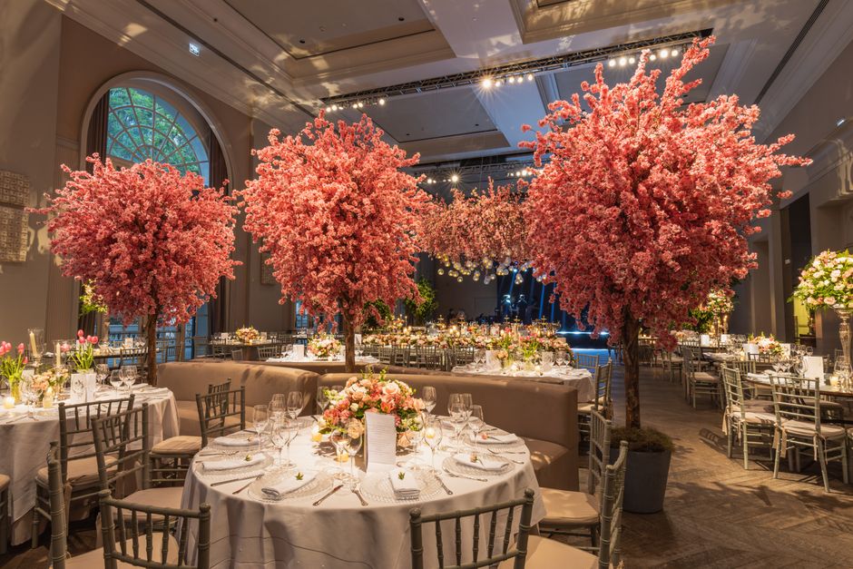 An opulent event hall decorated with multiple large pink cherry blossom trees with dense clusters of delicate petals, creating a lush, springtime ambiance. The tables are covered with white tablecloth