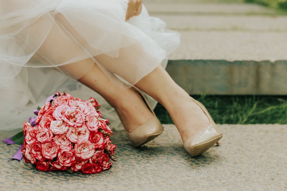 A close-up of a bride's legs and feet, showing her wearing beige high-heeled shoes and a flowing white wedding dress with tulle layers. She is seated on a stone bench outdoors, with a large bouquet of