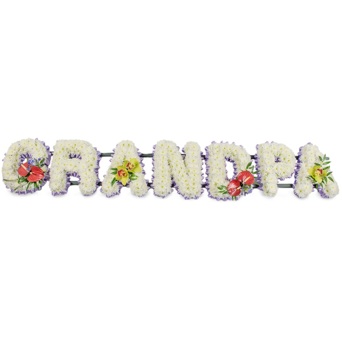 The photograph presents an elegant, bespoke funeral tribute spelling out "GRANDPA" in large, upright block letters, laid out in a straight horizontal line as if ready to be placed at the front of a service in Holborn. Each letter is meticulously covered in dense clusters of white chrysanthemums, the tightly layered petals creating a full, fluffy texture that gives the impression of soft cushions lined up side by side. The clear whiteness of the blooms suggests purity, remembrance, and a respectful farewell, their uniform shape and size lending the tribute a polished, professional finish. At the base of each floral letter, a narrow band of pale lavender ribbon forms a subtle edge, its gentle lilac hue offering just enough colour to trace the outline of the lettering without disturbing the calm, monochrome look. Hidden behind, slender dark green bars join the letters together, forming a single, robust structure that can be moved and displayed easily at a central London venue, from historic churches near Holborn Viaduct to modern crematoria within easy reach of the area. Four focal clusters of accent flowers interrupt the run of white. On the G, a compact arrangement of deep red anthuriums stands out, the glossy, heart-shaped spathes and bright yellow spadices contrasting beautifully with dark green foliage. The first A is adorned with yellow-green orchids, their delicate, finely veined petals and arching stems nestled among fresh leaves, adding a hint of sophistication. The D repeats the red anthuriums, providing visual balance and echoing the theme of heartfelt love, while the final A on the right mirrors the first A with more orchid blooms. This carefully balanced "GRANDPA" tribute is the kind of personalised funeral lettering an expert florist in Holborn might create to help families honour a cherished grandfather with grace and quiet beauty.